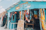 A colorful gift shop is displayed with an eclectic mix of items hanging and displayed outside. The exterior is painted bright blue, and there are many vibrant textiles, postcards, and tropical-themed decorations. The shop is open, revealing even more colorful goods inside.