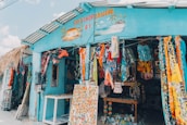 A colorful gift shop is displayed with an eclectic mix of items hanging and displayed outside. The exterior is painted bright blue, and there are many vibrant textiles, postcards, and tropical-themed decorations. The shop is open, revealing even more colorful goods inside.