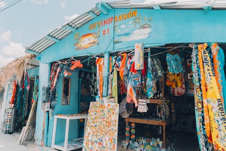 A colorful gift shop is displayed with an eclectic mix of items hanging and displayed outside. The exterior is painted bright blue, and there are many vibrant textiles, postcards, and tropical-themed decorations. The shop is open, revealing even more colorful goods inside.