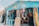 A colorful gift shop is displayed with an eclectic mix of items hanging and displayed outside. The exterior is painted bright blue, and there are many vibrant textiles, postcards, and tropical-themed decorations. The shop is open, revealing even more colorful goods inside.