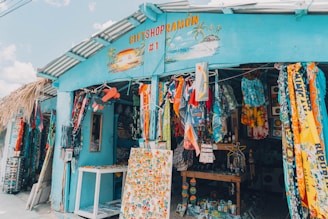 A colorful gift shop is displayed with an eclectic mix of items hanging and displayed outside. The exterior is painted bright blue, and there are many vibrant textiles, postcards, and tropical-themed decorations. The shop is open, revealing even more colorful goods inside.