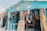 A colorful gift shop is displayed with an eclectic mix of items hanging and displayed outside. The exterior is painted bright blue, and there are many vibrant textiles, postcards, and tropical-themed decorations. The shop is open, revealing even more colorful goods inside.