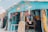 A colorful gift shop is displayed with an eclectic mix of items hanging and displayed outside. The exterior is painted bright blue, and there are many vibrant textiles, postcards, and tropical-themed decorations. The shop is open, revealing even more colorful goods inside.