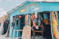 A colorful gift shop is displayed with an eclectic mix of items hanging and displayed outside. The exterior is painted bright blue, and there are many vibrant textiles, postcards, and tropical-themed decorations. The shop is open, revealing even more colorful goods inside.