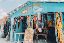 A colorful gift shop is displayed with an eclectic mix of items hanging and displayed outside. The exterior is painted bright blue, and there are many vibrant textiles, postcards, and tropical-themed decorations. The shop is open, revealing even more colorful goods inside.