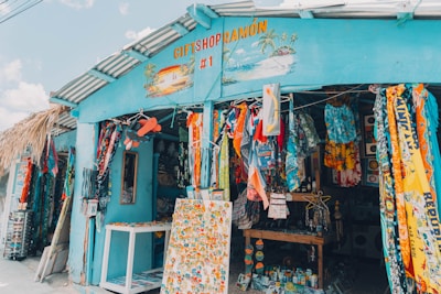 A colorful gift shop is displayed with an eclectic mix of items hanging and displayed outside. The exterior is painted bright blue, and there are many vibrant textiles, postcards, and tropical-themed decorations. The shop is open, revealing even more colorful goods inside.