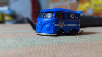 A small, blue toy van with stylish decals including a Union Jack and a retro racing number parked on a textured surface. The background is blurred, suggesting an indoor setting.