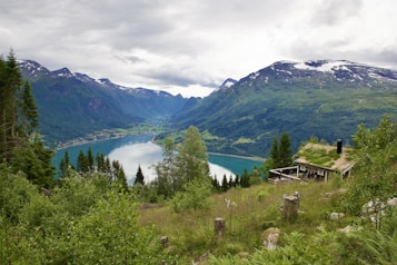 A picturesque view of a mountainous landscape with a serene lake surrounded by lush greenery. Snow-capped peaks rise in the background under a cloudy sky. A quaint wooden cabin with a grass-covered roof sits on the hillside, enveloped by trees and wild vegetation.