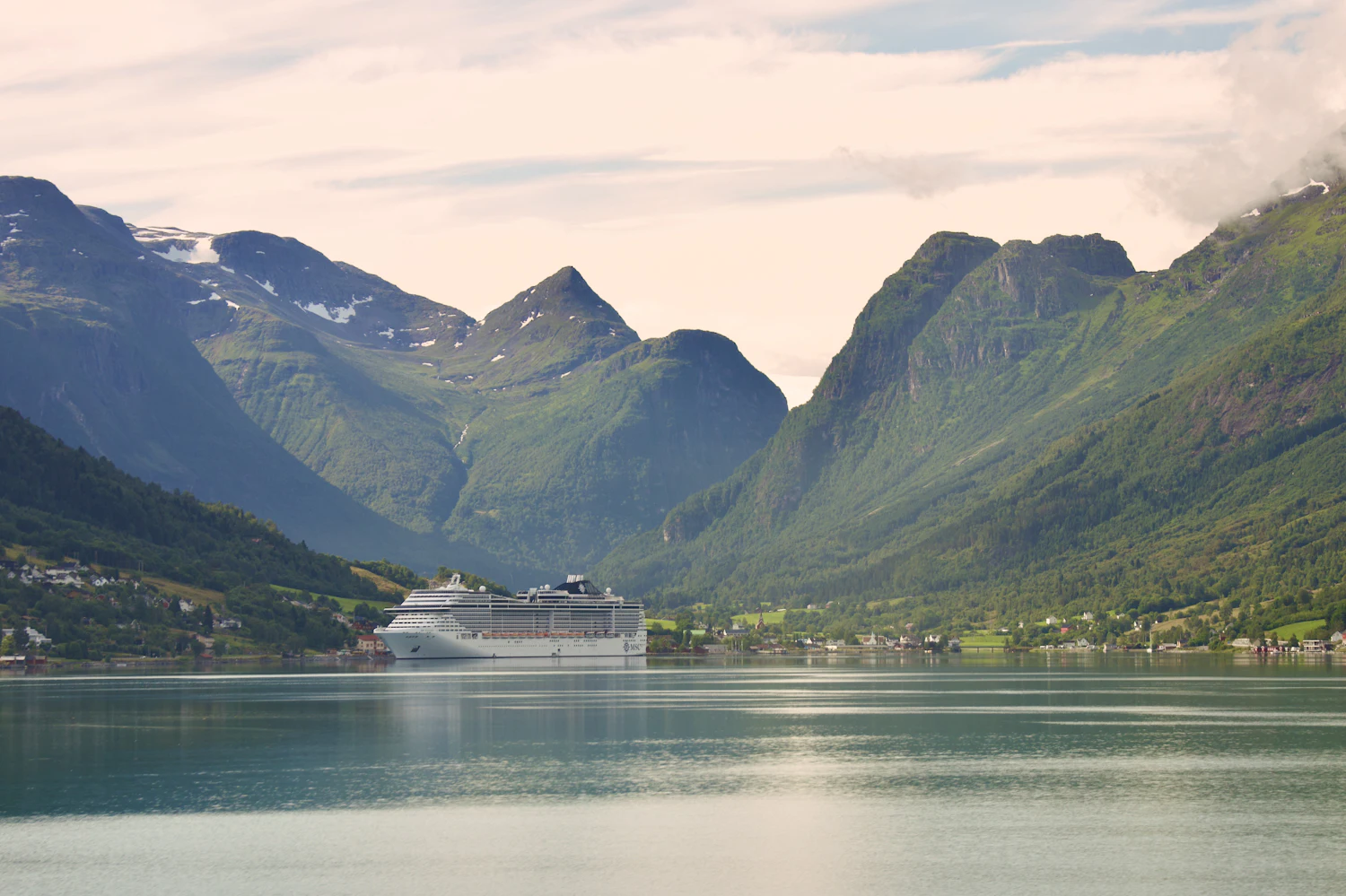 A single cruise ship at anchor in a wide, misty Norwegian fjord ringed by green mountains.