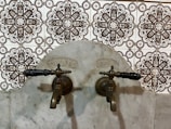Antique bronze taps mounted on a marble wall with intricate patterned tiles in the background. The design of the tiles is ornate, featuring symmetrical floral motifs in dark brown and white.