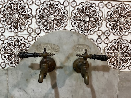 Antique bronze taps mounted on a marble wall with intricate patterned tiles in the background. The design of the tiles is ornate, featuring symmetrical floral motifs in dark brown and white.