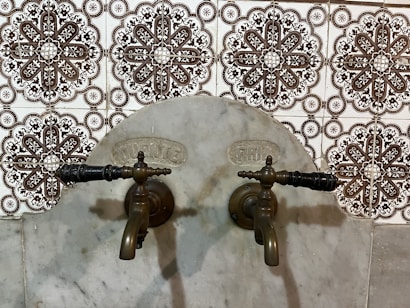 Antique bronze taps mounted on a marble wall with intricate patterned tiles in the background. The design of the tiles is ornate, featuring symmetrical floral motifs in dark brown and white.