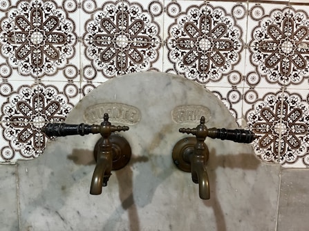 Antique bronze taps mounted on a marble wall with intricate patterned tiles in the background. The design of the tiles is ornate, featuring symmetrical floral motifs in dark brown and white.
