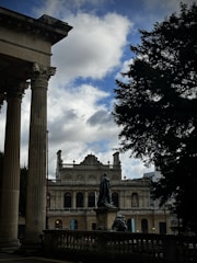 An architectural scene with classical elements, featuring tall columns in the foreground and a historic building with ornate details in the background. A bronze statue flanked by a railing is visible in the center, with a large tree on the right under a partly cloudy sky.