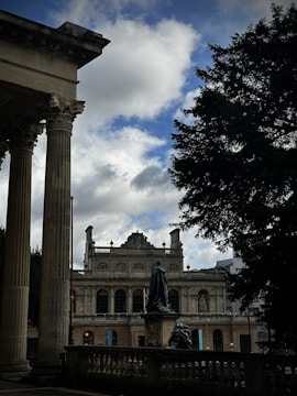 An architectural scene with classical elements, featuring tall columns in the foreground and a historic building with ornate details in the background. A bronze statue flanked by a railing is visible in the center, with a large tree on the right under a partly cloudy sky.