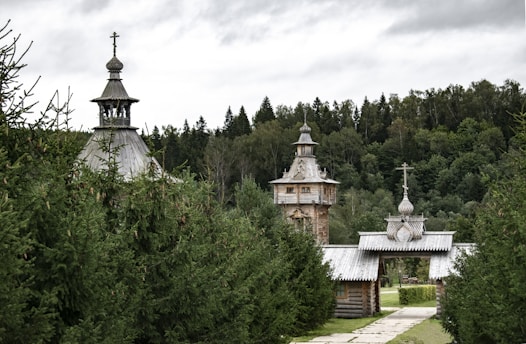 A serene landscape featuring traditional wooden architecture with ornate domes and crosses. The structures are surrounded by dense, lush green forest. A stone path leads towards the buildings, creating a sense of calm and seclusion.