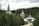 A serene landscape featuring traditional wooden architecture with ornate domes and crosses. The structures are surrounded by dense, lush green forest. A stone path leads towards the buildings, creating a sense of calm and seclusion.
