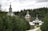 A serene landscape featuring traditional wooden architecture with ornate domes and crosses. The structures are surrounded by dense, lush green forest. A stone path leads towards the buildings, creating a sense of calm and seclusion.