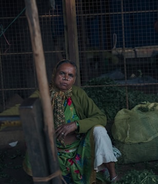 An elderly woman sits in a dimly lit space with a somber expression. She is wearing a green shawl and a colorful sari, adorned with a patterned scarf around her head. Next to her is a large sack, possibly containing produce. The background includes a metal grid fence, adding an industrial ambiance.