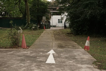 A paved pathway leads through a green, leafy area with traffic cones placed on either side. The path is marked with a white arrow pointing forward. In the background, a small building with a white facade and a few visible windows is partially obscured by trees.