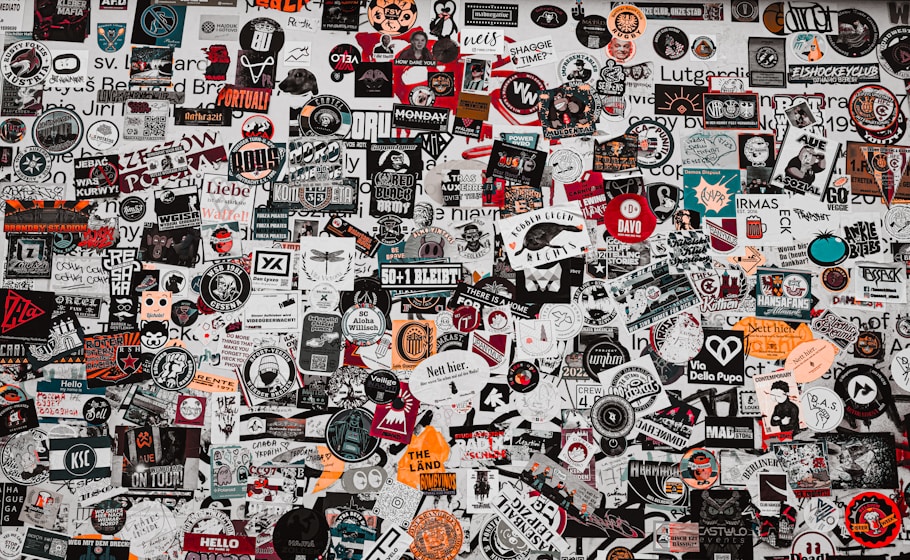 A dense collage of various stickers and logos covering a surface. The stickers include a mix of typography, graphics, and symbols, with some featuring slogans and brand names. The arrangement is chaotic, with different sizes, colors, and orientations overlapping each other.