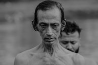 Two men are captured in a close-up portrait, one in front with a contemplative expression and the other blurred in the background, suggesting a moment of reflection or ritual by the water.