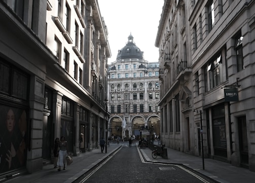 A narrow city street flanked by historic buildings featuring intricate architectural details. At the end of the street, a large building with a domed roof and multiple windows stands prominently. People are walking along the sidewalks, and signage for a store named 'Amalfi' is visible on the right. The scene likely takes place during the day, though lighting suggests a dim or overcast atmosphere.