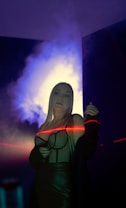 A person with long blonde hair stands in a dimly lit room with a dramatic, colorful glow around them. They are wearing a dark outfit with a visible structured bodice. A red laser light creates a line across their body, adding a dynamic element to the scene. The background features a soft, smoky texture with blue and purple hues.