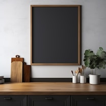A minimalist kitchen setup featuring a wooden countertop with cutting boards leaning against the wall, a collection of kitchen utensils in white holders, a potted plant with green leaves, and a large blank chalkboard with a wooden frame mounted on a light, textured wall.