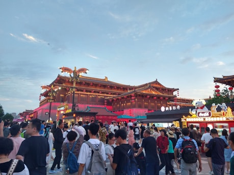 A bustling scene with a crowd of people walking and gathering in an outdoor area featuring traditional Chinese architecture. Red lanterns and ornate decorations adorn the large wooden structures. Food stalls or shops with bright signage are visible, adding to the lively atmosphere.