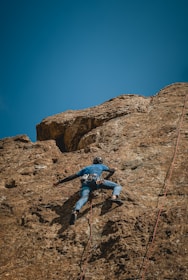 A person wearing climbing gear is ascending a steep, rocky cliff under a clear blue sky. The climber is secured with ropes and is focused on making their way up the uneven surface of the rock.