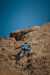 A person wearing climbing gear is ascending a steep, rocky cliff under a clear blue sky. The climber is secured with ropes and is focused on making their way up the uneven surface of the rock.
