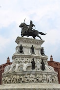 A grand monument featuring a detailed sculpture of a warrior on horseback at the top. Below are several tiers with carved figures, including soldiers and animals such as elephants, crafted in white stone. The structure is surrounded by architectural elements resembling fort walls.