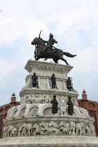 A grand monument featuring a detailed sculpture of a warrior on horseback at the top. Below are several tiers with carved figures, including soldiers and animals such as elephants, crafted in white stone. The structure is surrounded by architectural elements resembling fort walls.