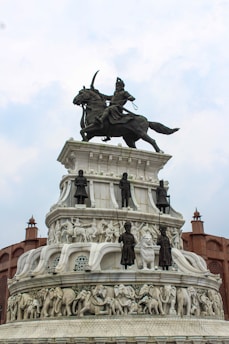 A grand monument featuring a detailed sculpture of a warrior on horseback at the top. Below are several tiers with carved figures, including soldiers and animals such as elephants, crafted in white stone. The structure is surrounded by architectural elements resembling fort walls.