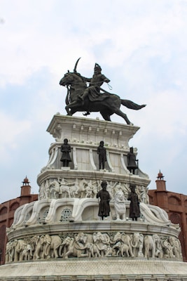 A grand monument featuring a detailed sculpture of a warrior on horseback at the top. Below are several tiers with carved figures, including soldiers and animals such as elephants, crafted in white stone. The structure is surrounded by architectural elements resembling fort walls.