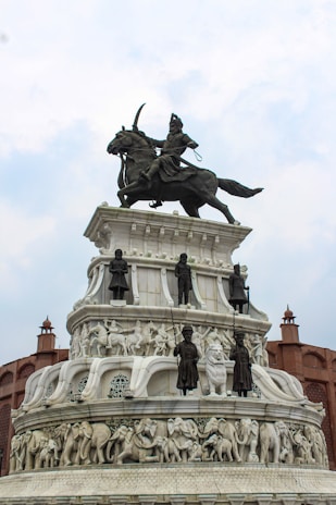A grand monument featuring a detailed sculpture of a warrior on horseback at the top. Below are several tiers with carved figures, including soldiers and animals such as elephants, crafted in white stone. The structure is surrounded by architectural elements resembling fort walls.