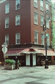 A corner street scene featuring a three-story red brick building with several windows. The ground floor houses a restaurant named Sevilla, with an awning over the entrance and outdoor seating. Street signs and a decorative streetlamp are also visible alongside some potted plants.