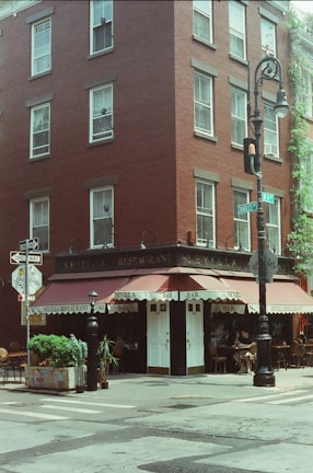A corner street scene featuring a three-story red brick building with several windows. The ground floor houses a restaurant named Sevilla, with an awning over the entrance and outdoor seating. Street signs and a decorative streetlamp are also visible alongside some potted plants.