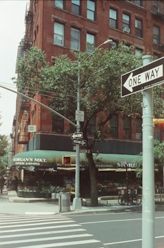 A busy urban street corner features a brick building with several windows and a classic fire escape. A green awning displaying 'Morgan's Mkt. Open 24hrs' covers a grocery market with visible fresh produce and flowers. A large tree partially obscures the building, while multiple street signs, including a one-way sign, are clearly visible. Bicycles are parked by the curb, and a traffic light is overhead.
