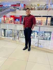 A person in a maroon shirt and dark trousers is standing in a modern shopping mall. The background features various stores with colorful signboards, including clothing and accessories shops. The setting is lively with other shoppers visible in the background and an escalator to the left.