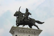 A bronze statue of a warrior on horseback, mounted on a tall pedestal. The warrior holds a weapon aloft, wearing traditional armor and headgear. The horse is in a dynamic pose, adding to the sense of movement. The sky is overcast, providing a neutral backdrop.