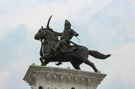 A bronze statue of a warrior on horseback, mounted on a tall pedestal. The warrior holds a weapon aloft, wearing traditional armor and headgear. The horse is in a dynamic pose, adding to the sense of movement. The sky is overcast, providing a neutral backdrop.