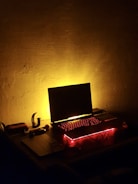 A dimly lit room with a laptop on a desk, surrounded by red and yellow ambient lighting that casts a warm glow. The laptop's keyboard is illuminated with red backlighting, sitting next to a few small objects, possibly electronic devices and accessories.