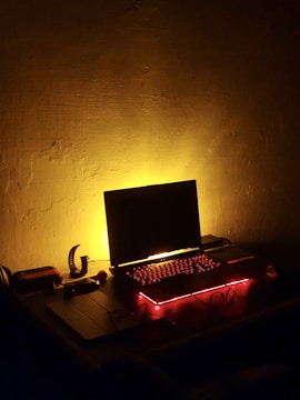 A dimly lit room with a laptop on a desk, surrounded by red and yellow ambient lighting that casts a warm glow. The laptop's keyboard is illuminated with red backlighting, sitting next to a few small objects, possibly electronic devices and accessories.