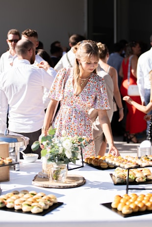A young woman in a floral dress is reaching for food at a buffet table during an outdoor event. The table is adorned with platters of small sandwiches and rolls, with a centerpiece of a floral arrangement. People in the background, dressed in casual to semi-formal attire, are socializing under bright sunlight.