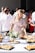 A young woman in a floral dress is reaching for food at a buffet table during an outdoor event. The table is adorned with platters of small sandwiches and rolls, with a centerpiece of a floral arrangement. People in the background, dressed in casual to semi-formal attire, are socializing under bright sunlight.
