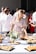 A young woman in a floral dress is reaching for food at a buffet table during an outdoor event. The table is adorned with platters of small sandwiches and rolls, with a centerpiece of a floral arrangement. People in the background, dressed in casual to semi-formal attire, are socializing under bright sunlight.