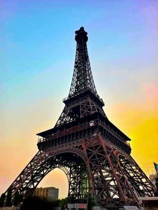A view of the Eiffel Tower during sunset, with a gradient sky transitioning from blue to shades of yellow and orange. The intricate iron lattice structure is silhouetted against the colorful backdrop, providing a striking contrast.