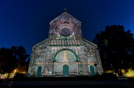 A historic stone church facade is illuminated in the night sky, showcasing intricate architectural details like a circular stained glass window and arched entryways. The lighting casts colorful patterns on the stone surface, contrasting with the deep blue of the sky. Trees frame the church on either side, adding a natural element to the scene.